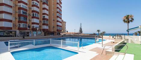 A swimming pool with a white fence, a palm tree, and a building with balconies in the background.