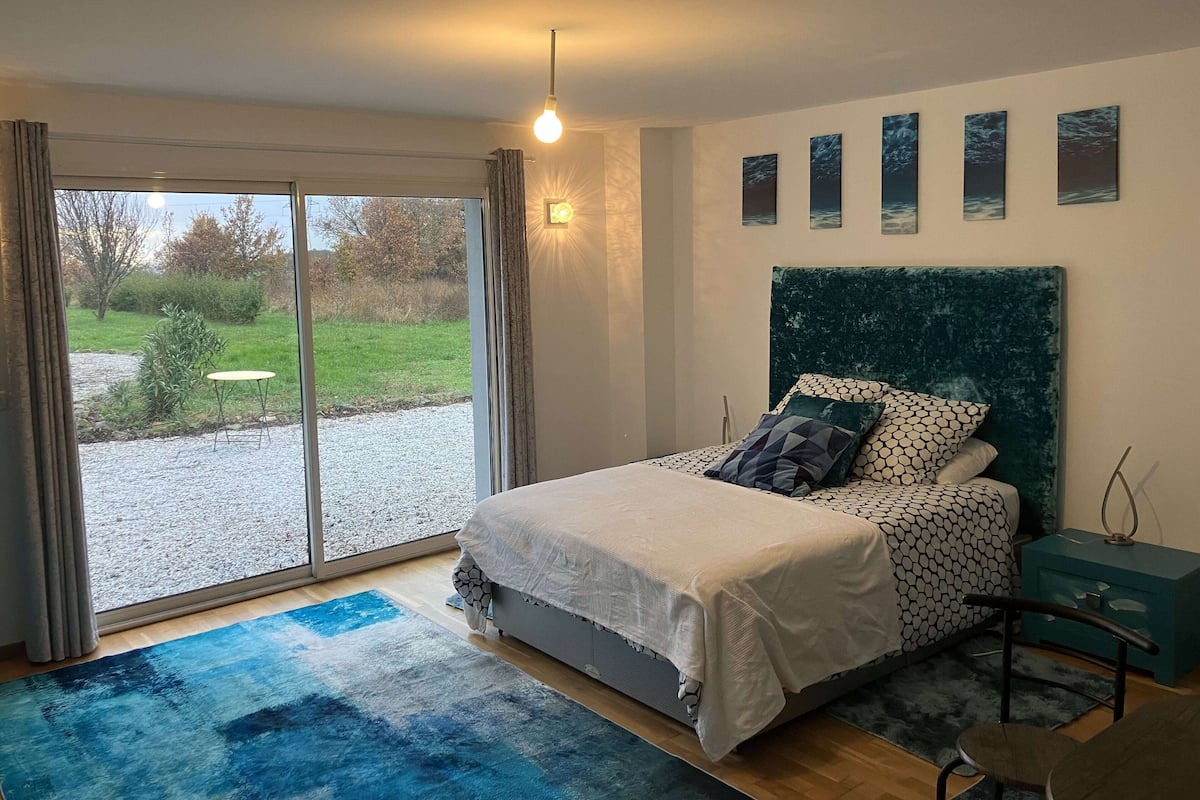 A bedroom with a bed, a nightstand, a chair, a blue rug, and a view of a garden through a sliding door.