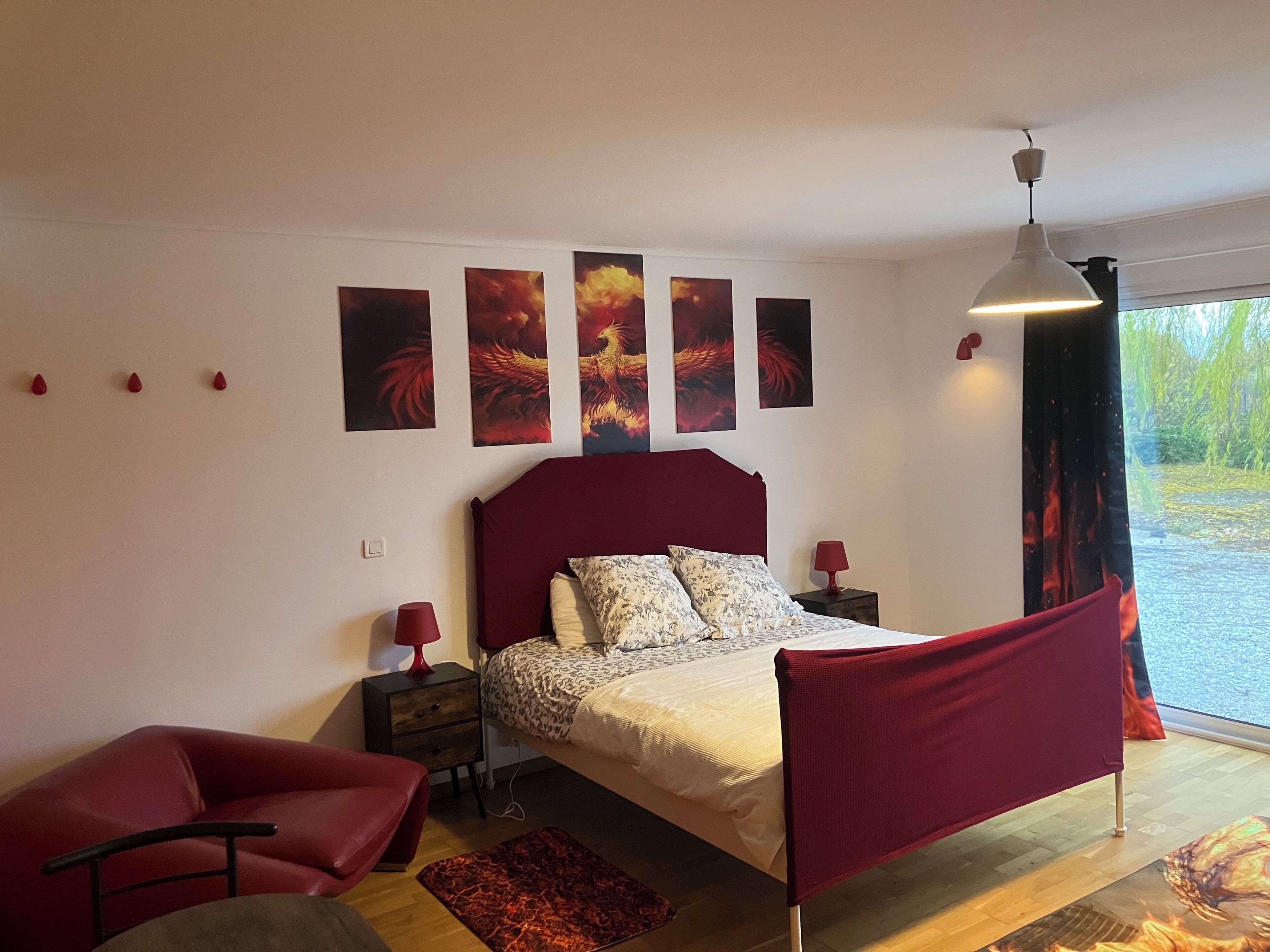 A bedroom with a red headboard, a bed with floral bedding, a red chair, a nightstand with a lamp, and a window with a view of greenery.