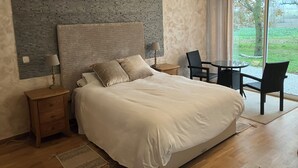 A bedroom with a large bed, a nightstand, a lamp, a chair, a small table, and a view of the outdoors through a window. - Chez PhiL (La Pomarède)