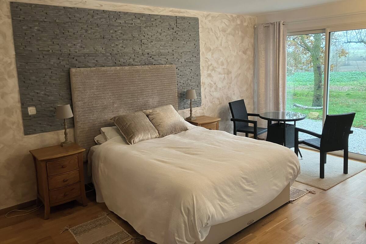 A bedroom with a large bed, a nightstand, a lamp, a chair, a small table, and a view of the outdoors through a window.