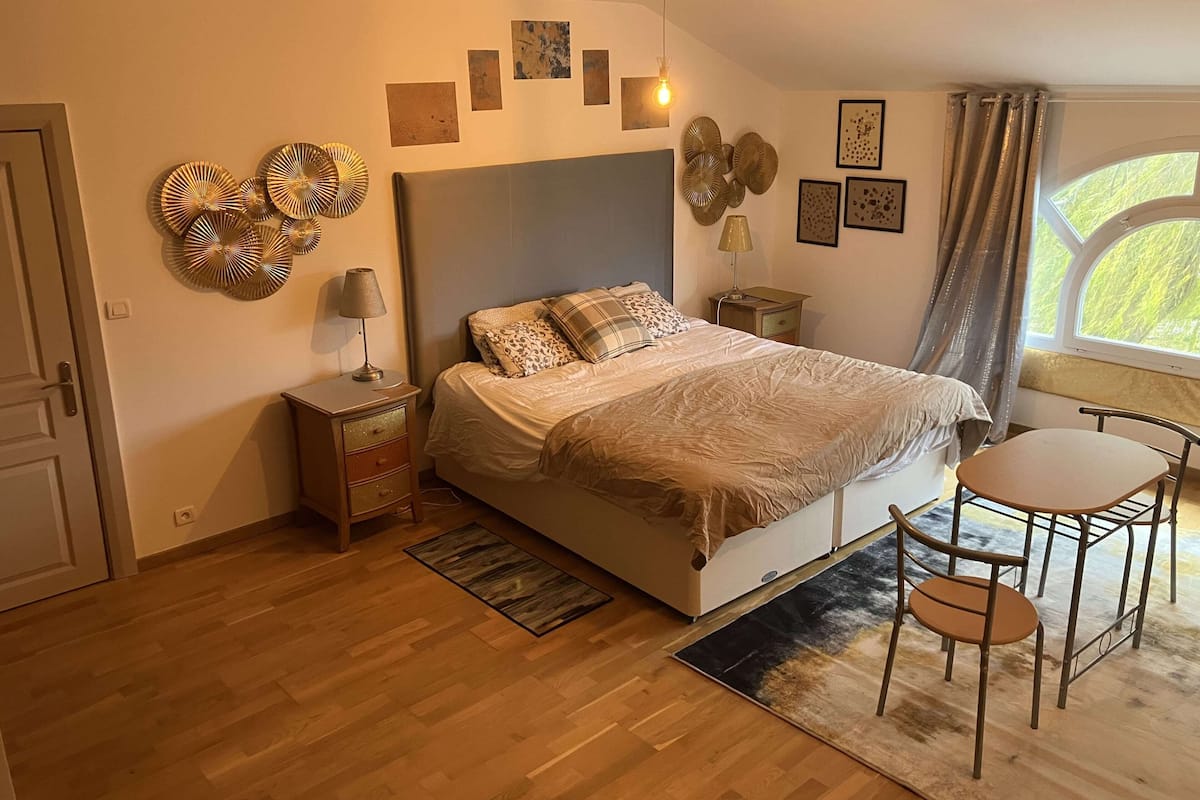 A bedroom with a bed, bedside table, lamp, decorative wall art, and a window with curtains.