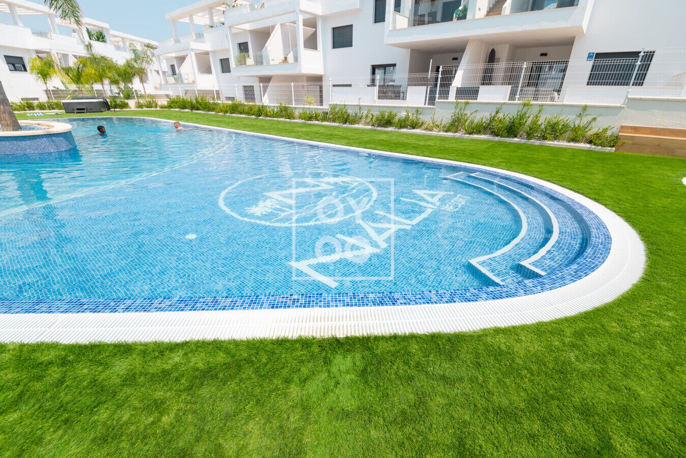 A modern swimming pool with a logo, surrounded by well-maintained grass and white buildings.
