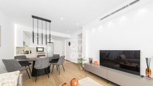 A modern living room with a flat-screen TV, grey sofas, a dining area with a table and chairs, and a kitchen in the background. - Nalia Horizon Penthouse, Outdoor living with solarium, BBQ, pool, hot tub - by OYOOSTAYS (Torrevieja)