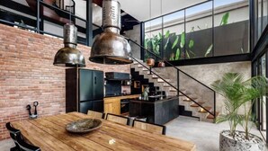 A modern kitchen with a brick wall, wooden dining table, and a large window with a plant. - Villa Koma by BaliSuperHost (Bali)