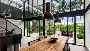 A modern kitchen with a wooden table and hanging pendant lights, overlooking a lush garden with palm trees. - Villa Koma by BaliSuperHost (Bali)