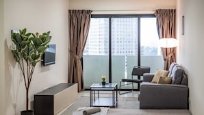 A modern living room with a sofa, a coffee table, a TV, a potted plant, and a balcony with curtains. - A Lovely & Comfy Suite at Suasana Suites JB 11 (Johor Bahru)