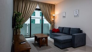 Un salon moderne avec un canapé gris, une table basse en bois et un téléviseur posé sur un meuble. On y trouve une grande fenêtre avec des rideaux, une plante et un coussin rouge sur le canapé.
