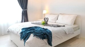 Un dormitorio moderno con una cama blanca, cortinas grises, una lámpara de noche y una planta pequeña en la mesita.