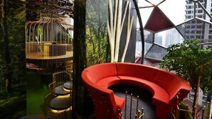 Un intérieur moderne avec un canapé rouge aux lignes courbes, un escalier en colimaçon et une vue sur le paysage urbain, encadrée par une structure de fenêtre aux motifs géométriques.