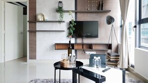 Un salon moderne avec un téléviseur à écran plat fixé à un mur en bois, une table basse ronde agrémentée d’objets décoratifs et une grande fenêtre avec des rideaux.
