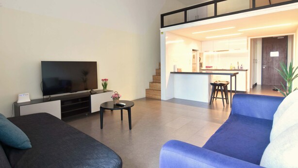 A modern living room with a flat-screen TV, a sofa, a coffee table, and a blue armchair. A kitchen is visible in the background with a dining area and a staircase leading to an upper level. - Loft in Prime Bangsar Location (Kuala Lumpur)