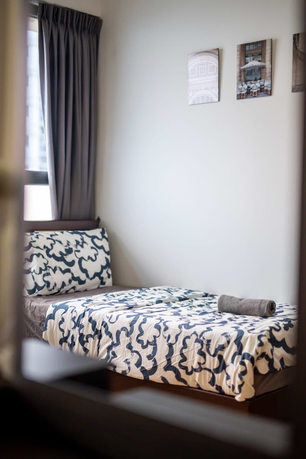 A small, neatly made bed with a patterned bedspread, a pillow, and a folded blanket on a wooden floor.
