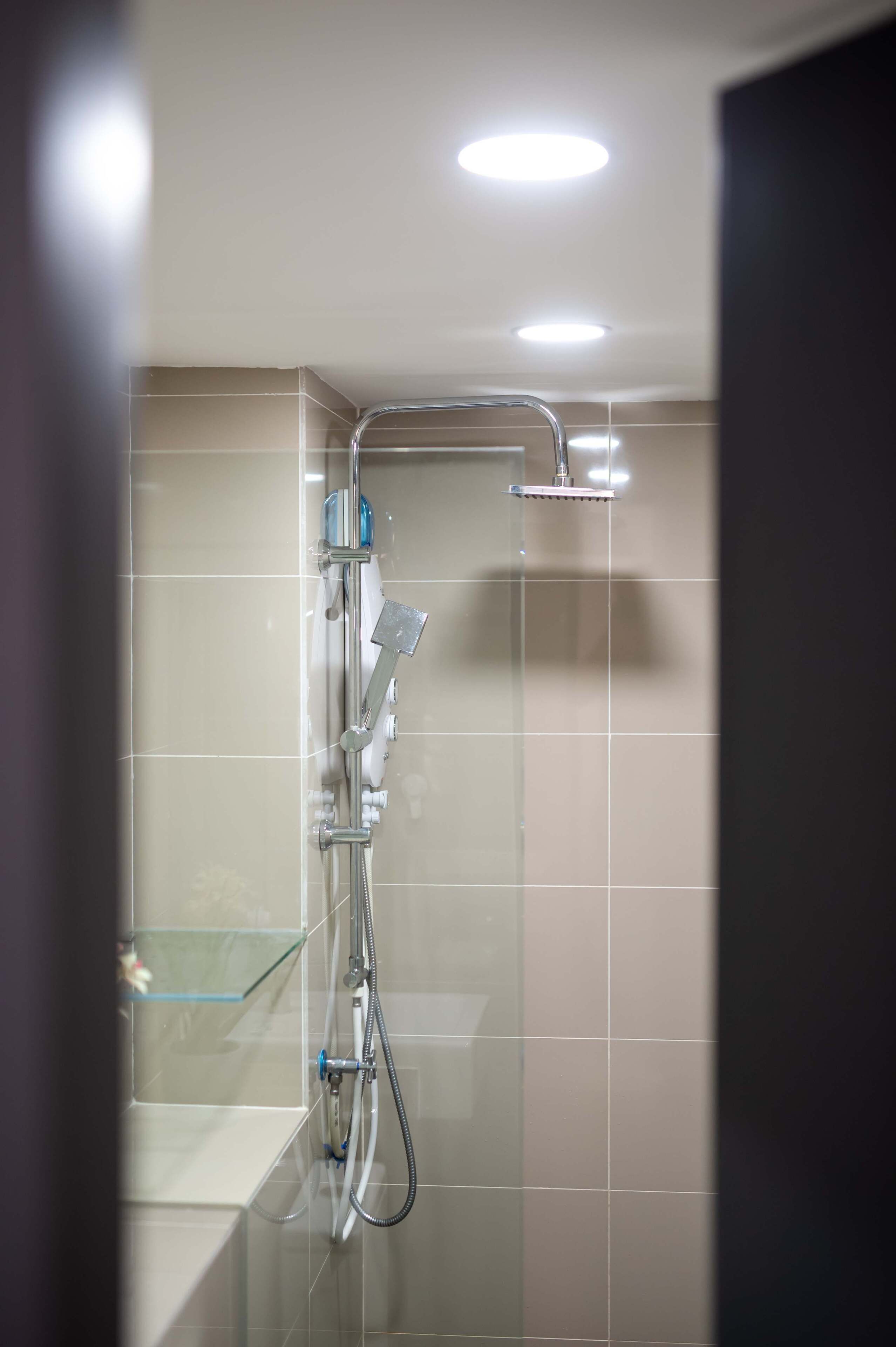 A shower with a handheld showerhead and a wall-mounted faucet, a glass shelf, and a tiled wall.