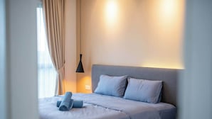 Una cama bien hecha con cabecera gris, una lámpara colgante negra y una ventana con cortinas.