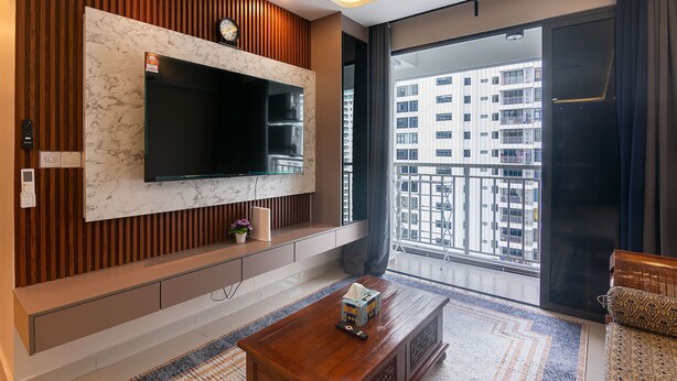 A modern living room with a large TV mounted on a marble wall, wooden paneling, a sofa, a coffee table, and a view of a high-rise building through a large window. - 3BR Lsh33 KL Duke Highway Genting Free parking (Kuala Lumpur)