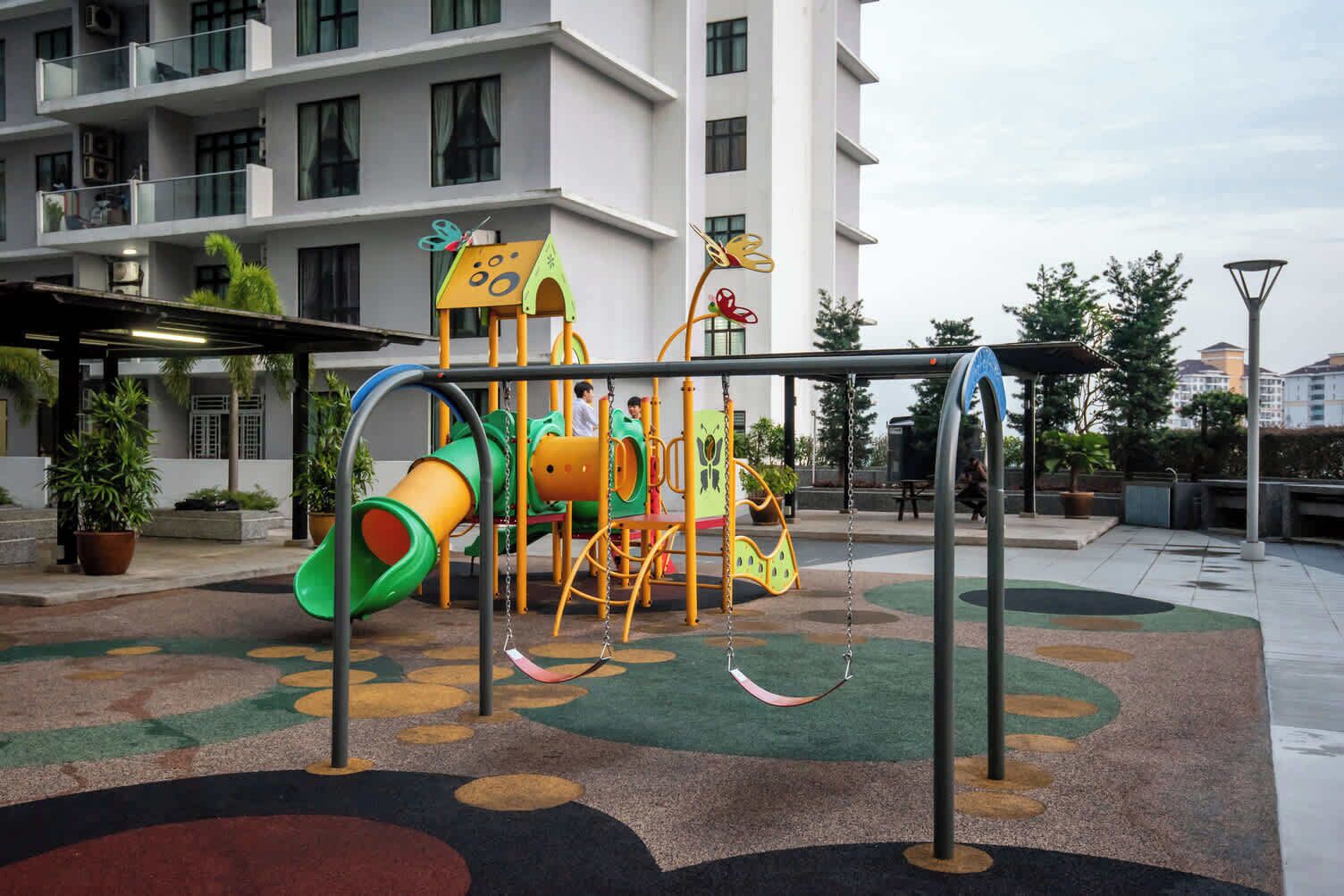 Un parque infantil con un tobogán y estructuras para escalar, rodeado de plantas en macetas y un edificio al fondo.