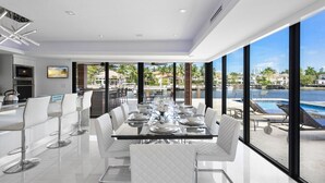 Un espace de restauration moderne avec une longue table dressée pour six personnes, de grandes fenêtres offrant une vue sur une piscine et sur le bord de l’eau.