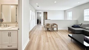 Un salon moderne avec un canapé, une table basse et un téléviseur. Il y a un coin cuisine équipé d’un évier et de placards. La pièce est dotée d’un plancher en bois et de murs blancs.