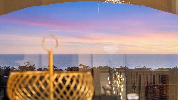 A view through a window with a decorative metal bowl in the foreground, a balcony railing, and a sunset over the ocean.