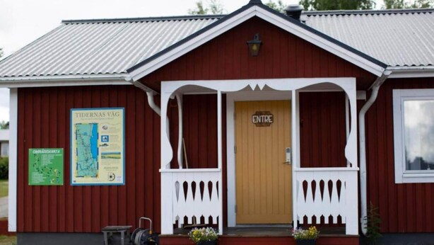 Un edificio rosso con una porta in legno recante la scritta "ENTRE", un portico con ringhiere bianche e due vasi di fiori sui gradini.