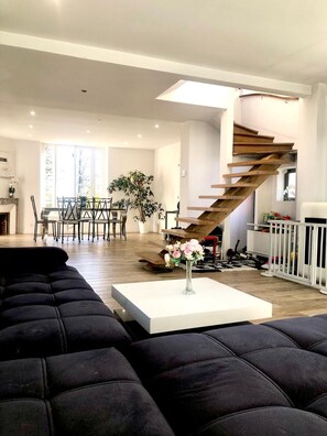Un salon moderne avec un canapé d’angle noir, une table basse blanche ornée d’un vase de fleurs, un coin repas avec une table et des chaises, et un escalier en bois.