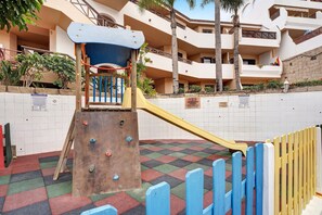 A playground with climbing structures and a slide, surrounded by a white wall and a multi-story building with balconies.