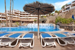 Un área de piscina con sillones de descanso, una sombrilla de paja y una pared de piedra.