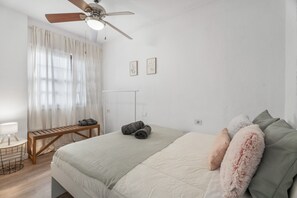 A bedroom with a bed, a wooden bench, a desk with a lamp, and a ceiling fan.