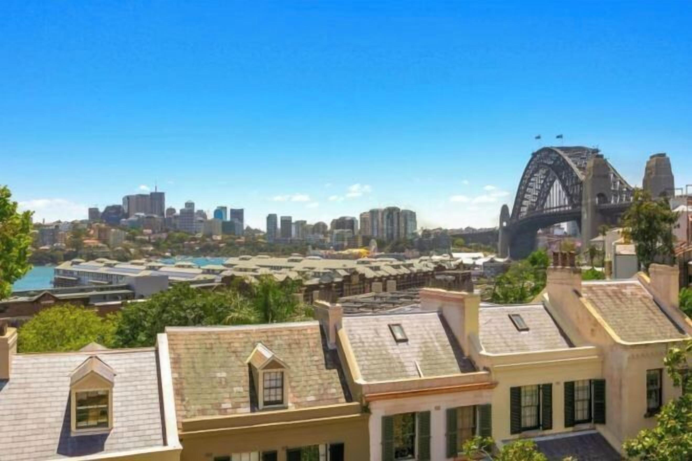 A coastal cityscape with a prominent bridge, likely the Sydney Harbour Bridge, and a cluster of buildings with flat roofs and green shutters.