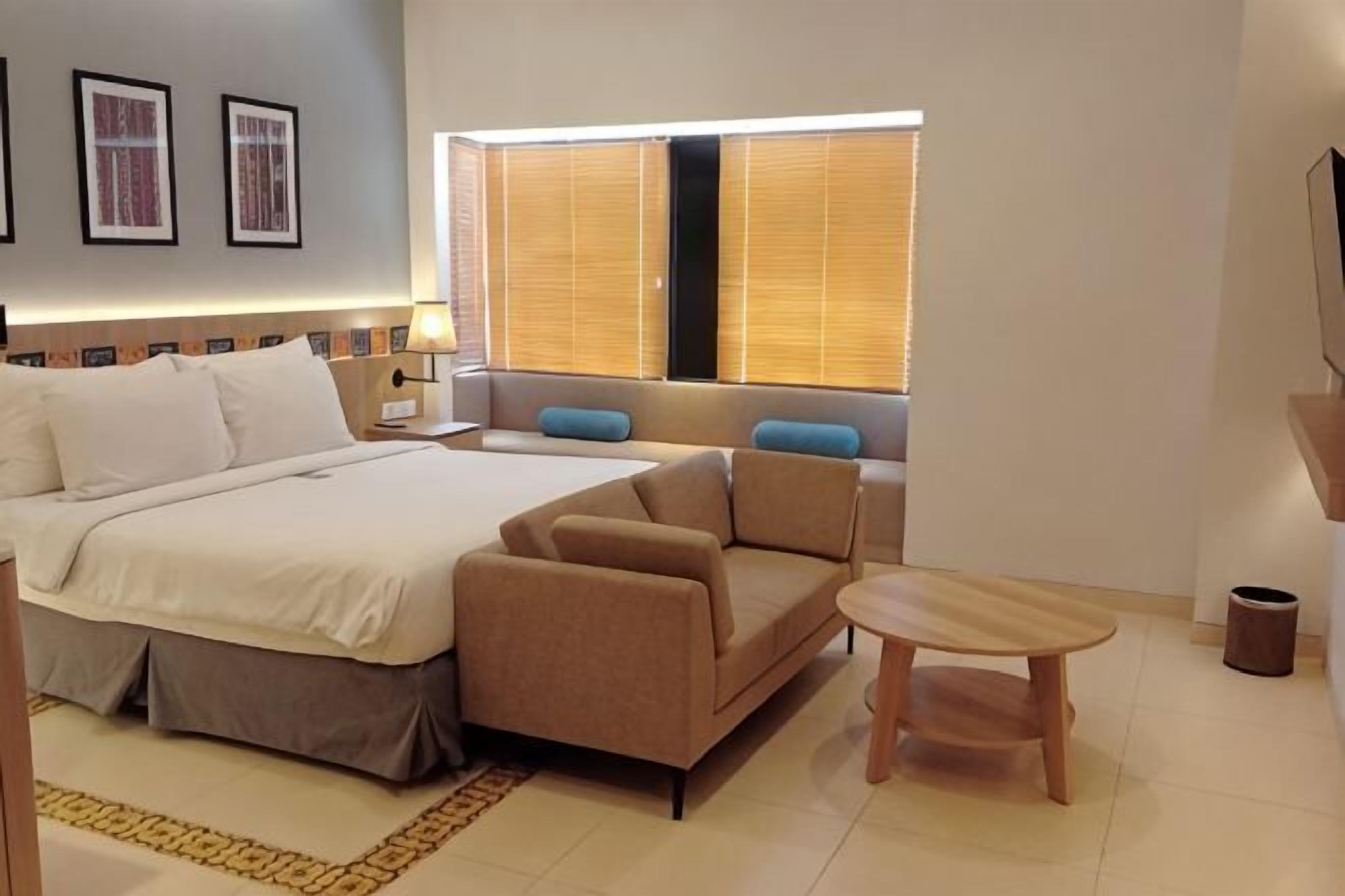 A hotel room with a bed, a sofa, a small table, a window with blinds, and a wall-mounted TV.