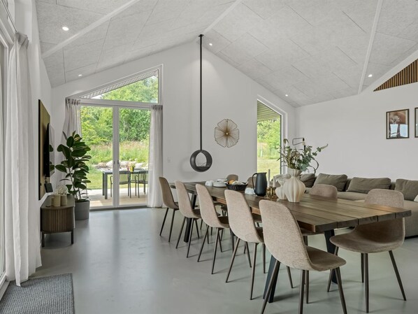 A spacious dining area with a long table, multiple chairs, a sofa, and a large window offering a view of greenery.