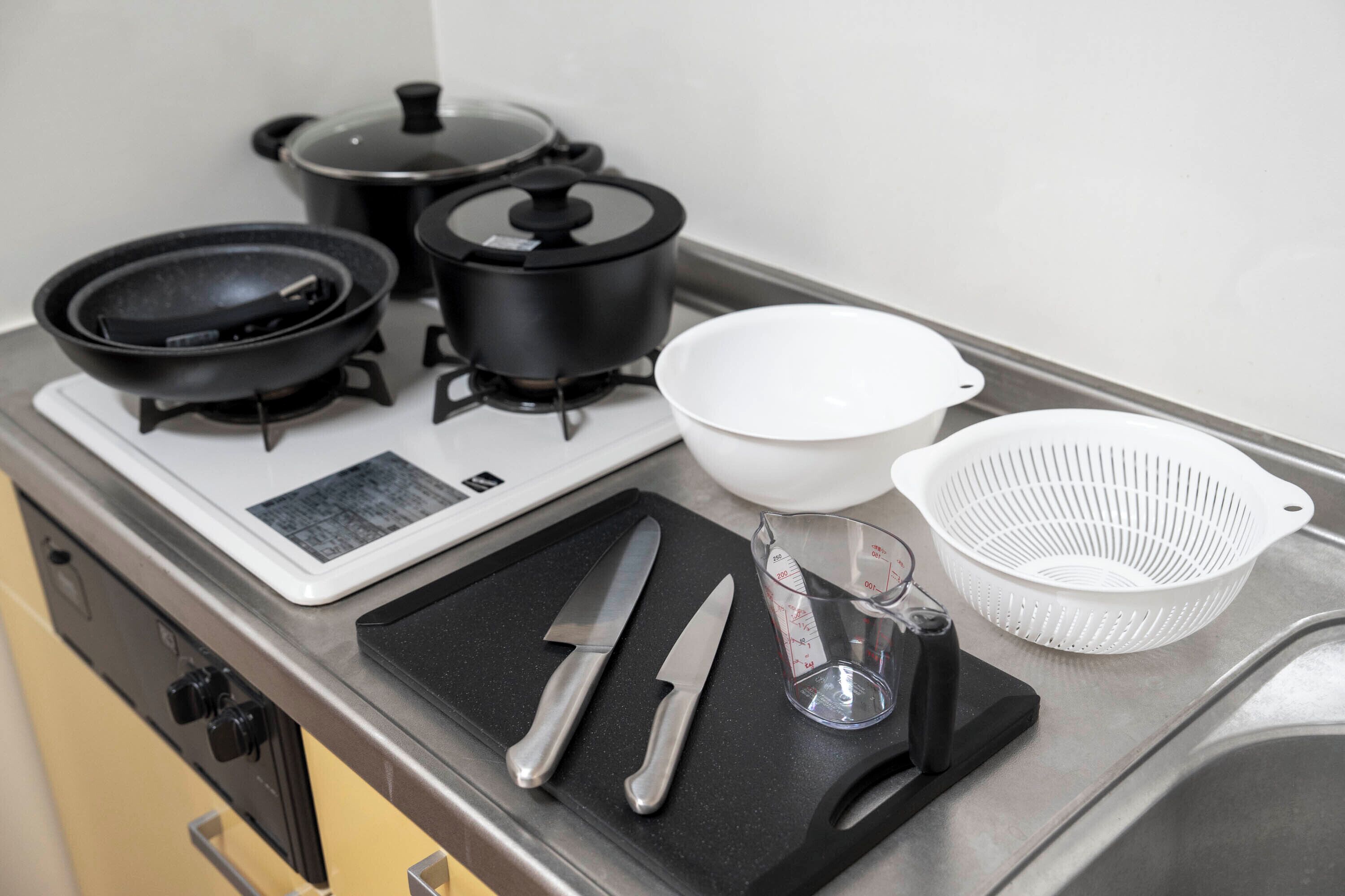 A kitchen counter with a stove, pots, a frying pan, a white bowl, a colander, a measuring cup, and two knives.