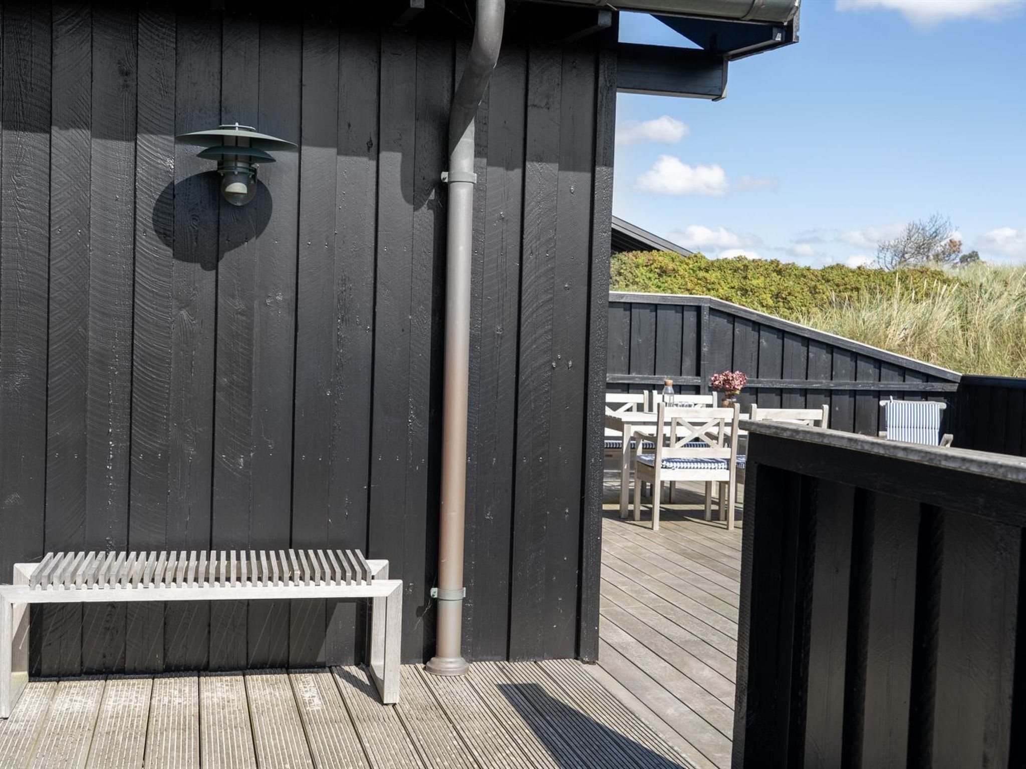 Une terrasse en bois avec un banc et une rambarde, un luminaire en métal et une vue sur un bâtiment doté d’une zone abritée.