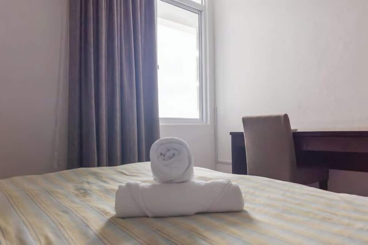 Hotellihuone, jossa on sänky, rullalle taitettu pyyhe, ikkuna verhoilla ja työpöytä tuolilla.