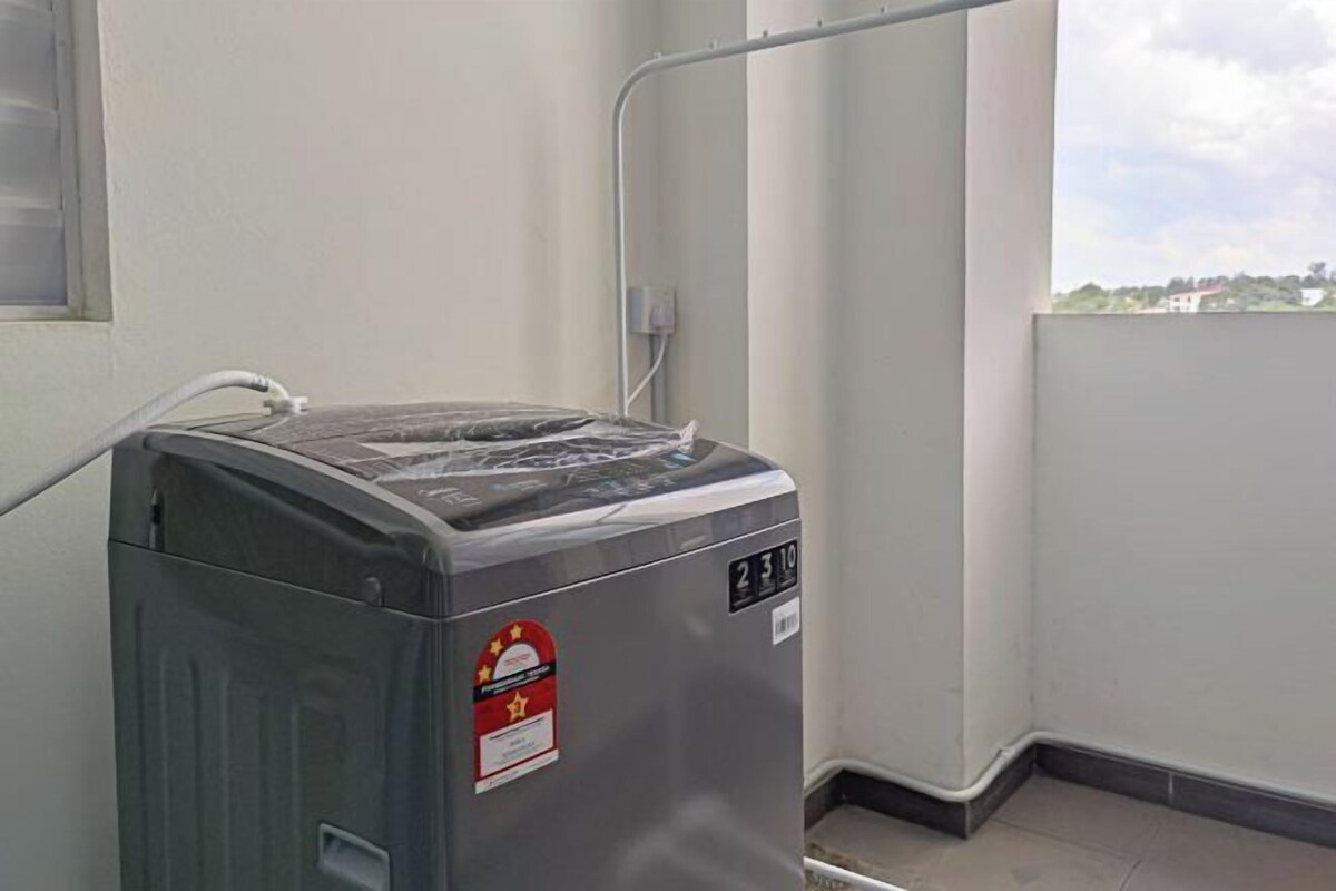 A compact, gray laundry machine with a water inlet hose, located in a corner of a room with a window and a white wall.