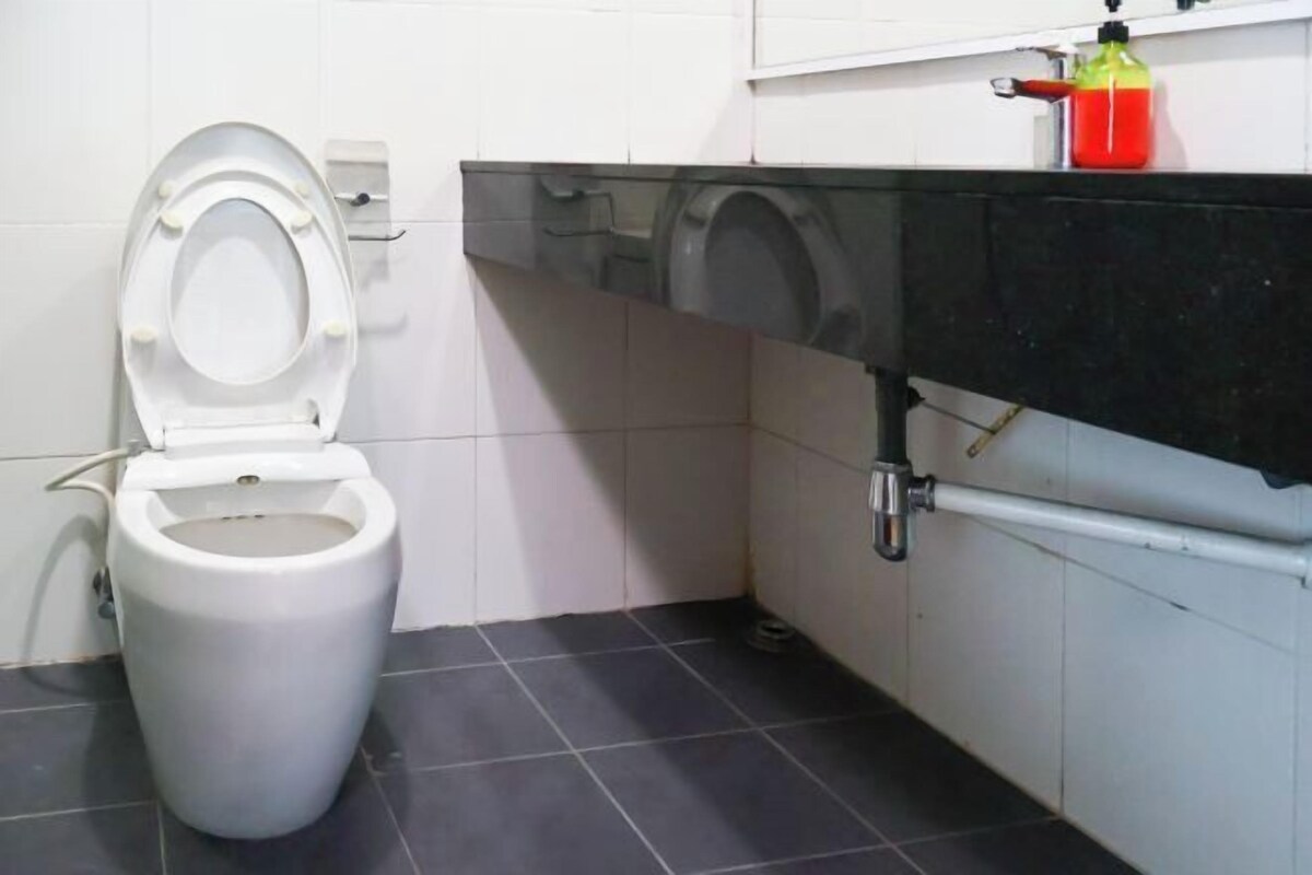 A toilet with a white seat and lid, a black tiled floor, a white wall, and a black countertop with a red bottle.