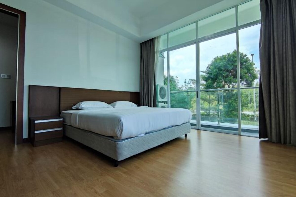 A bedroom with a large bed, wooden flooring, a nightstand, a window with curtains, and a view of greenery outside.