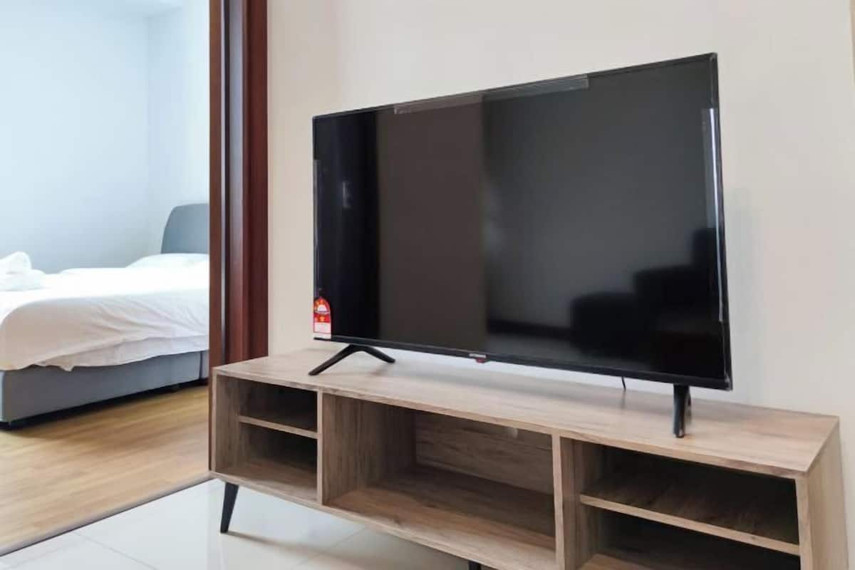 Moderni hotellihuone, jossa on suuri, litteänäyttöinen televisio puisen jalustan päällä, valkoisilla lakanoiden peittämä sänky ja näkyvillä oleva palosammutin.