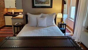 Quarto de hotel com cama, secretária pequena, cadeira, mesa de cabeceira, abajur e um quadro na parede.