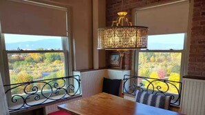 Uma área de refeições com uma mesa e cadeiras de madeira, uma luminária decorativa e vista da folhagem outonal através de grandes janelas.