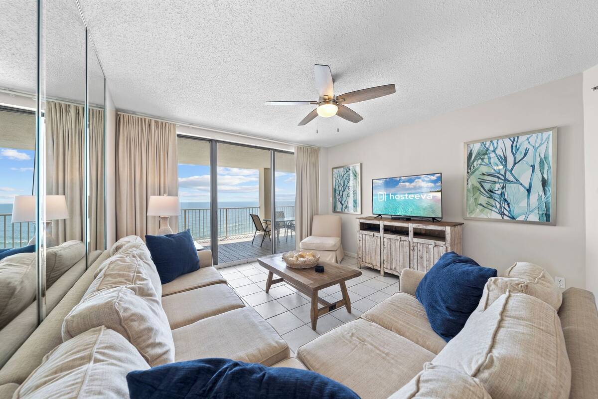 A spacious living area with a large sofa set, a wooden coffee table, a ceiling fan, a television, and artwork on the walls.