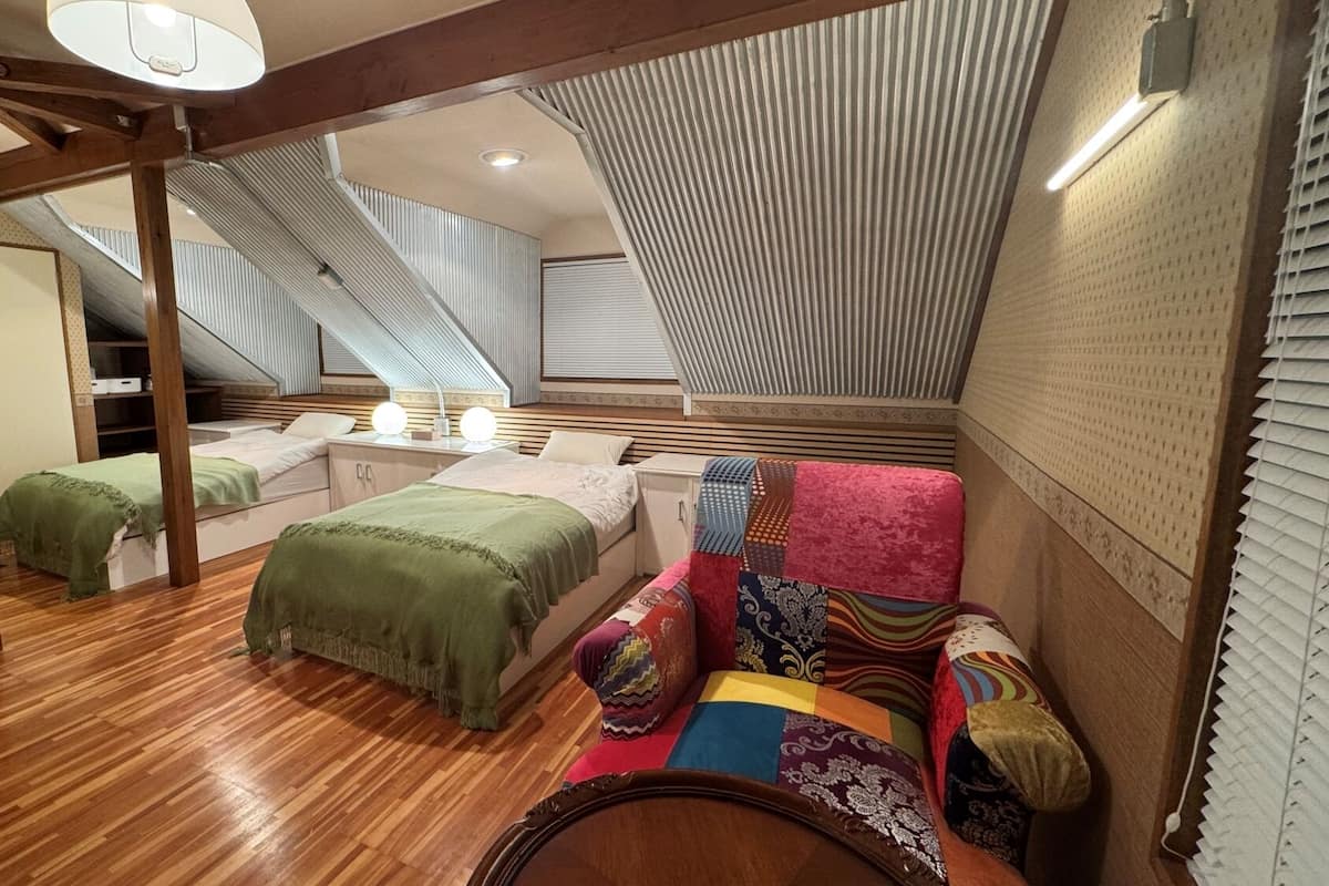 A bedroom with a sloped ceiling, wooden beams, a bunk bed, a sofa with colorful cushions, and a small table with lamps.