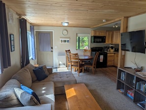 A cozy living room with a sofa, a coffee table, and a dining area with chairs and a table. There is a kitchen area with a microwave, stove, and cabinets. A TV is mounted on the wall, and a bookshelf is visible.