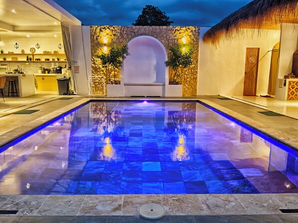 Una piscina con iluminación azul, un muro de piedra con plantas y una estructura con techo de paja.