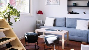 Un salon moderne avec un canapé gris, une table basse en bois et des chaises noires et blanches. On peut voir une lampe rouge, un tableau encadré et des étagères ornées d’objets décoratifs.