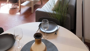 Un salon moderne avec un canapé, une table à manger pour deux personnes, une lampe sur pied et une grande fenêtre donnant sur l’extérieur.