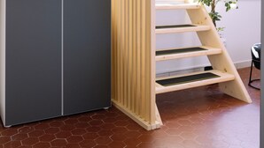 Une pièce avec une armoire grise, un escalier en bois et une grande fenêtre donnant sur des chaises placées à l’extérieur.