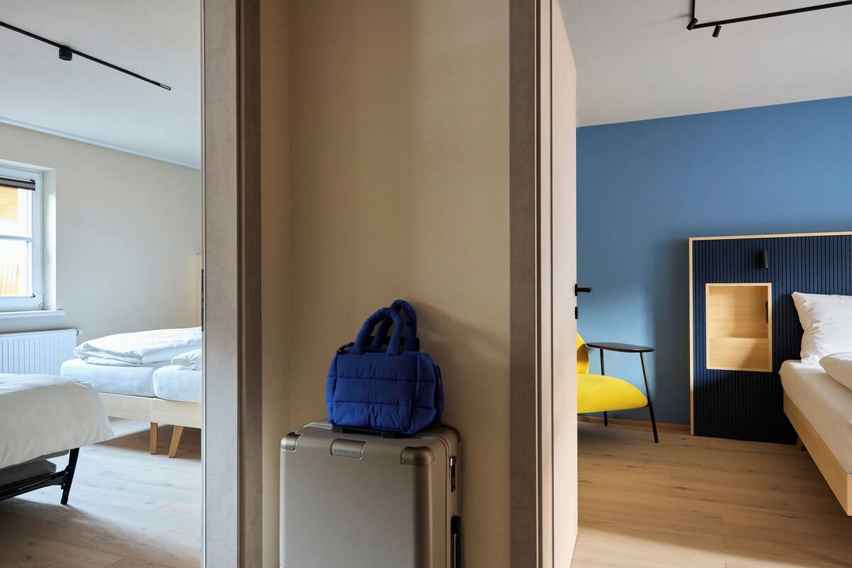 A hotel room with a bed, a suitcase, a blue bag, and a small table with a chair.