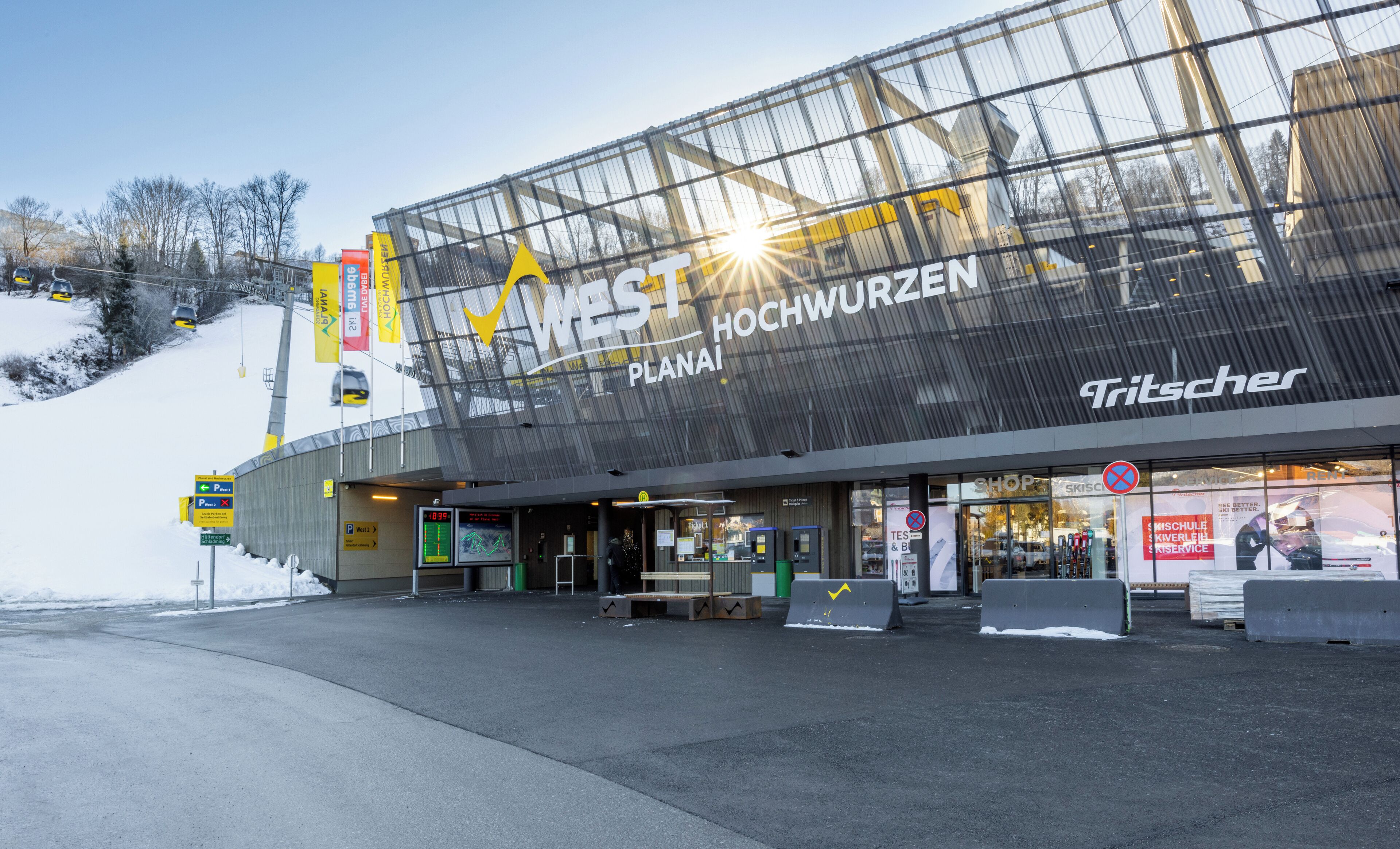 A modern building with a large glass facade, signage for 'WEST Hochwurz', and a 'Tritscher' shop.